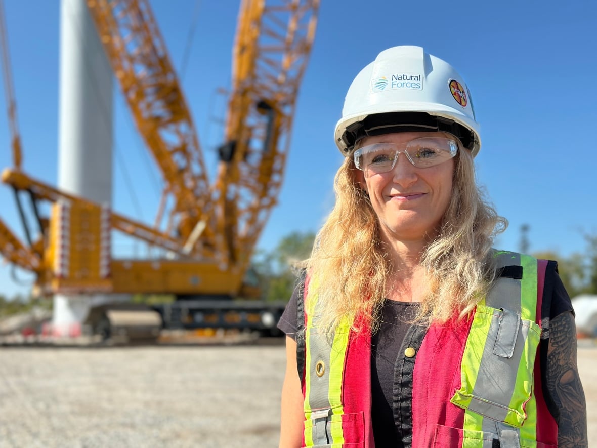 A woman in a hard hat and safety goggles smiles at the camera. The base of a crane is seen in the background.