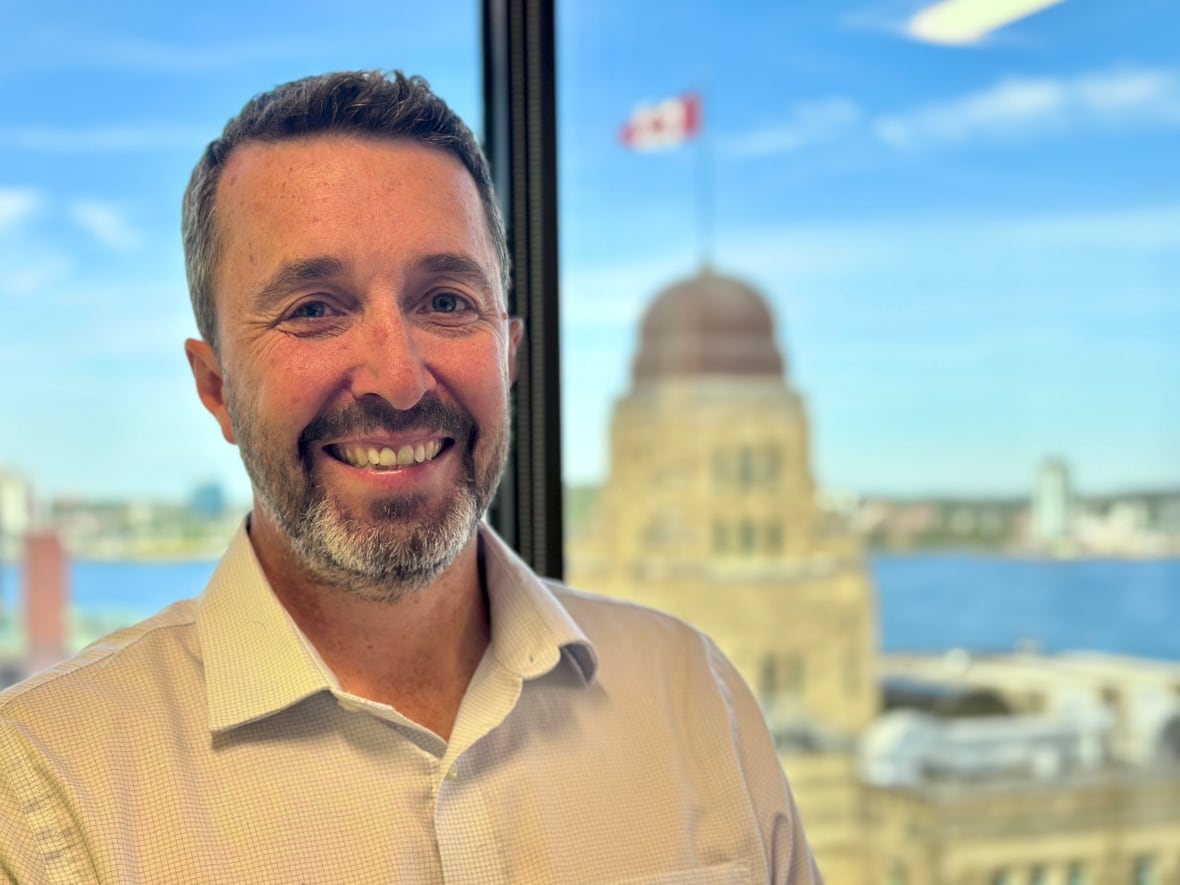 A man smiles at the camera. Parts of the downtown Halifax skyline and harbour are seen in the background through a window.
