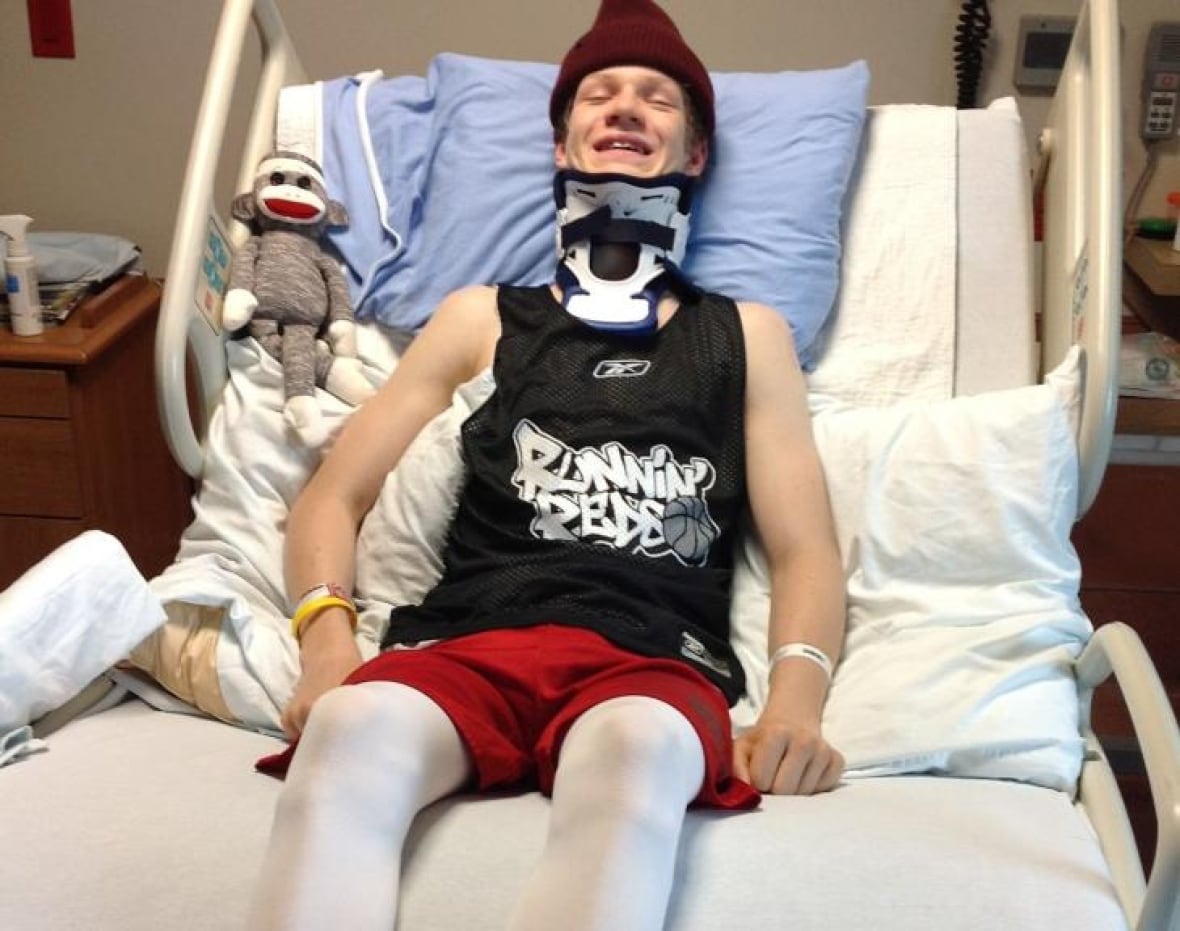 A teenage boy wearing a neck brace lays in a bed wearing a toque, shirt and shorts, with compression socks on his legs. He is propped up by pillows and smiling.