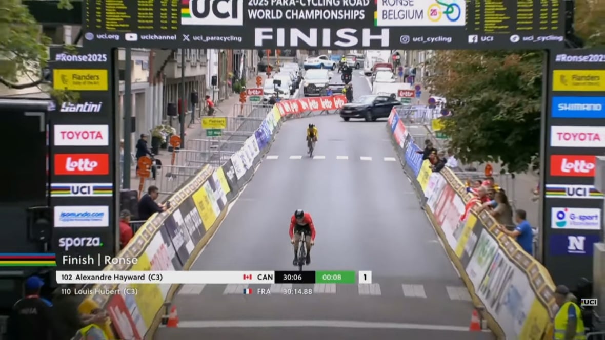 A wide shot of a man in a red jersey riding a bike across a finish line, with the name "Alexandre Hayward" and "30:06" on screen.
