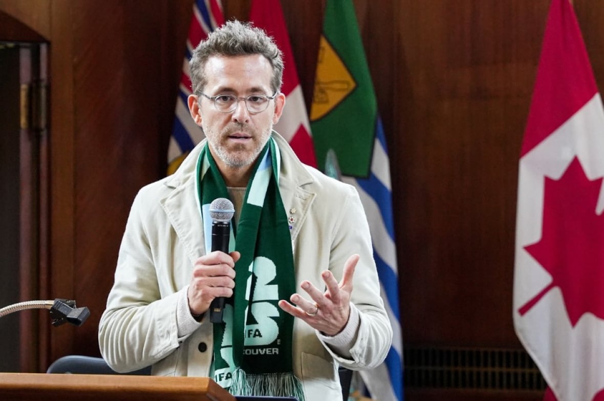 Ryan Reynolds, gekleed in een bruine blazer en een groene sjaal, spreekt op een podium.