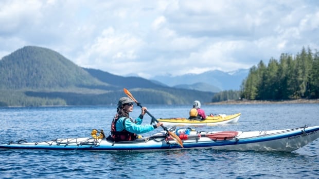 Photo shows two kayakers in water with forest and mountains behind them 
