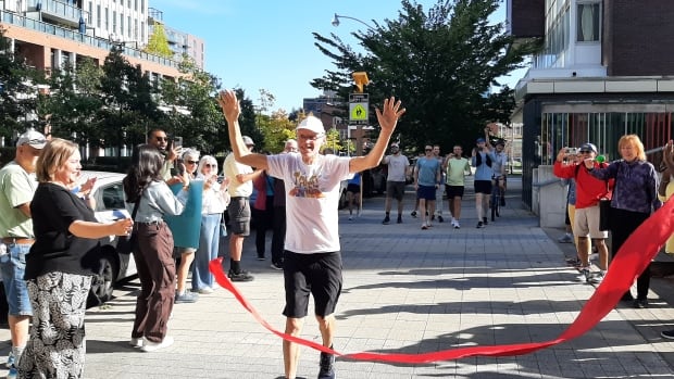 A man running on through a red ribbon on a city street sidewalk. He is smiling and has his hands in the air. Crowds are gathered on either side of the sidewalk, applauding and taking videos.