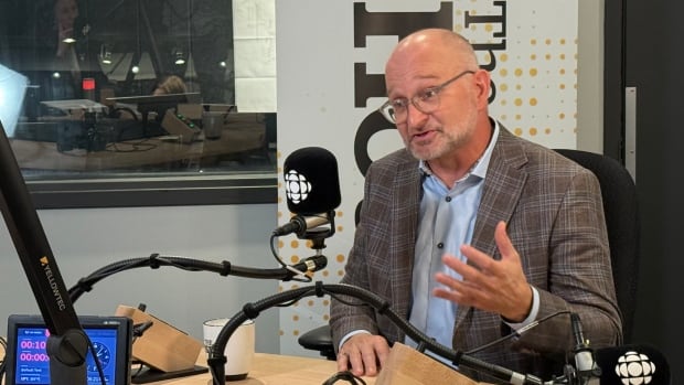 A bald man with a blue dress shirt and brown plaud jacket speaks into a CBC microphone