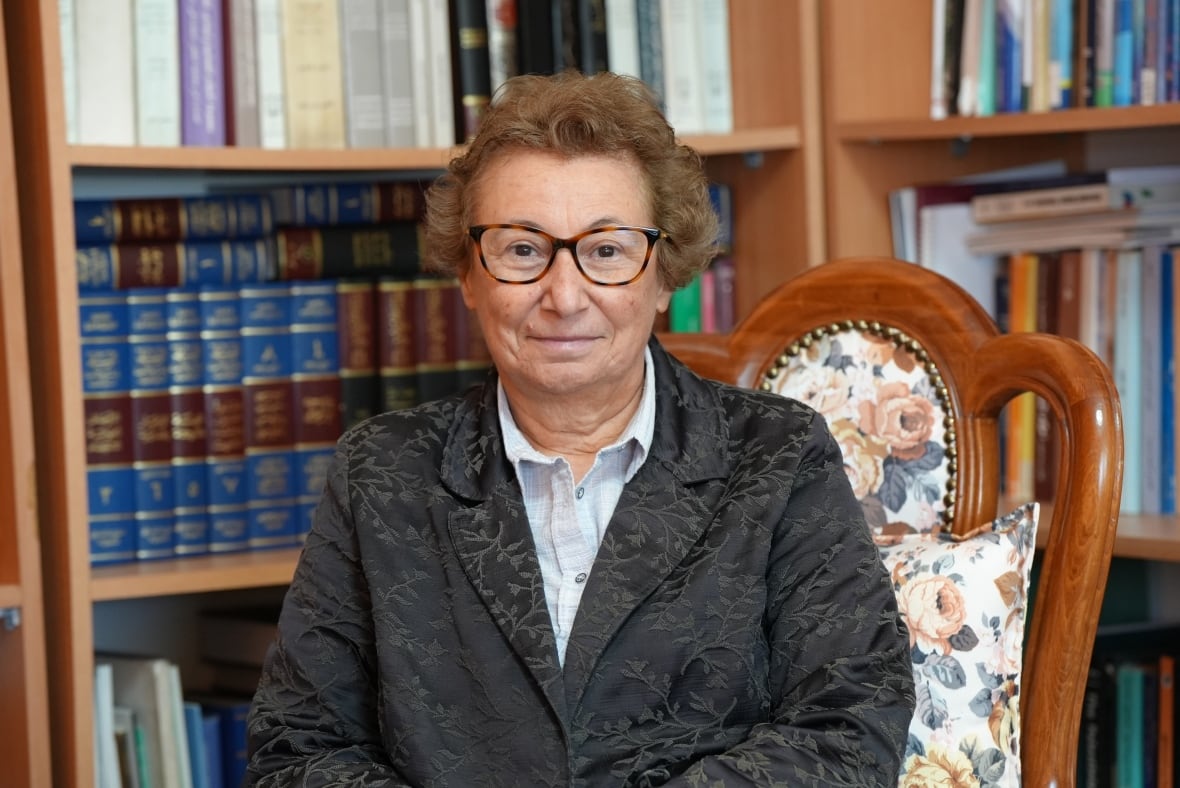 A woman in a black jacket sits on a chair. She wears brown glasses and has brown hair. Behind it was written on the shelves.