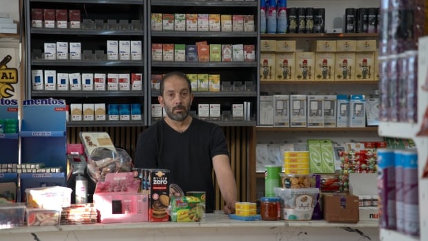 A man in a black V-neck stands behind the counter of a shop. Around him are various food products.