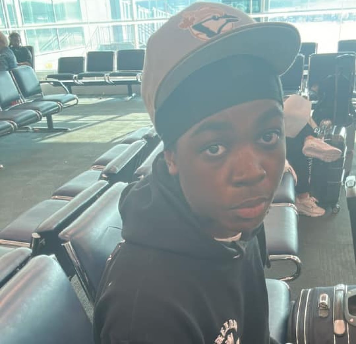 A Black boy with a Toronto Blue Jays hat on sits on a seat in an airport.