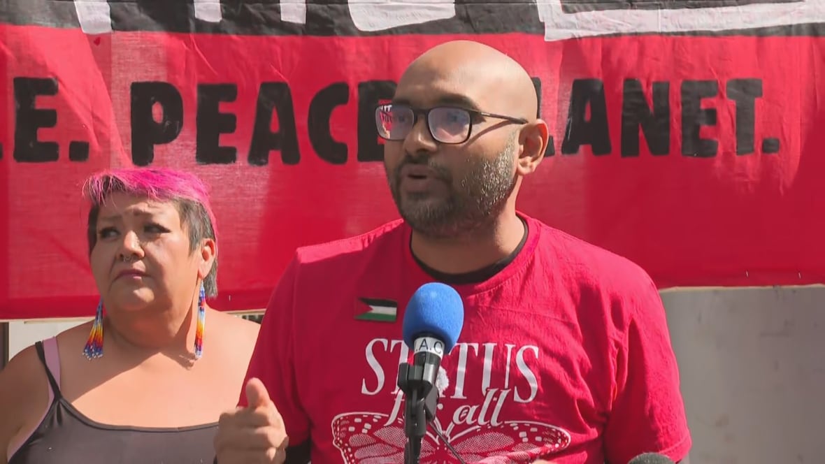 A middle aged bald man in glasses and a red shirt speaks into a microphone in front of a sign that reads "peace. planet." It is a sunny day outside. A woman is standing behind him.
