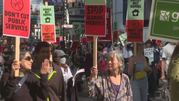 A large crowd of protesters holding signs about public service cuts, war and housing march through downtown Toronto on a sunny September afternoon