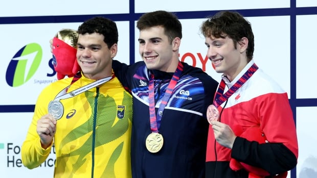 Three male Para swimmers smile while standing on a podium with medals around their necks.
