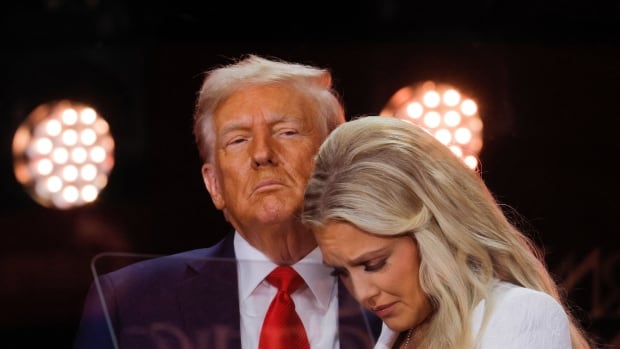 A woman looks down while standing in front of a man in a suit on a stage.
