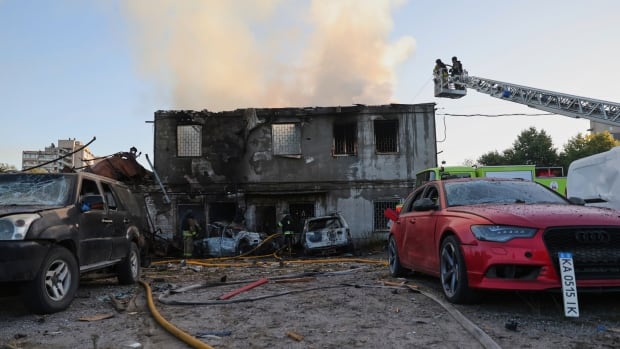 A two-storey residential building is seen destroyed and smoking, with firefighters in a bucket responding. Their are two vehicles in front of the house.