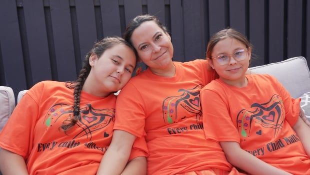 Melanie Cormier and sits in between her daughters. They are all wearing orange shirts that say Every Child Matters.