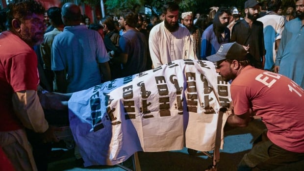 Several people gather around or help transport a body on a stretcher which is covered.