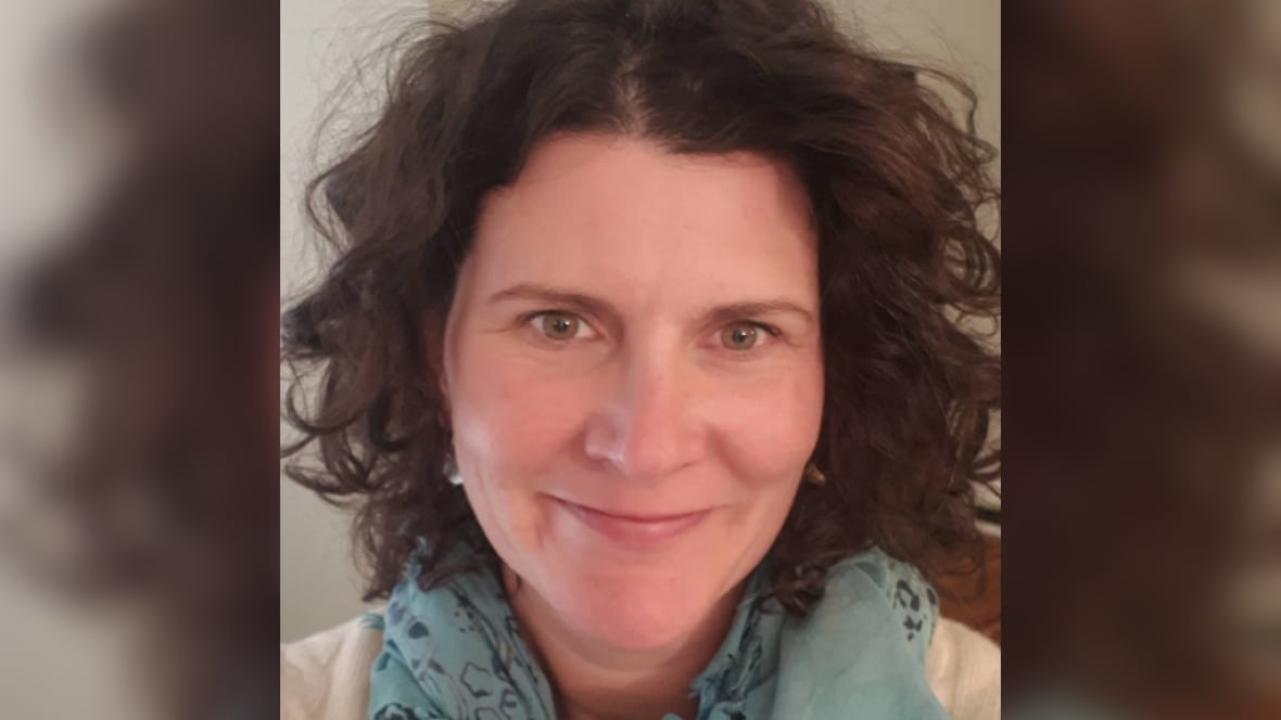 A woman with brown curly hair, wearing a light blue scarf with a dark blue pattern on it, smiles.