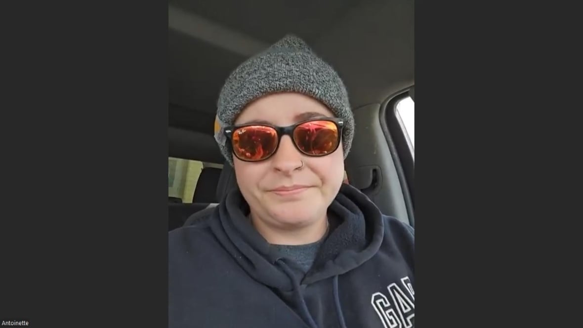 A woman in her car, wearing a grey toque, black sunglasses with red mirrored lenses, and a navy blue gap hoodie. 