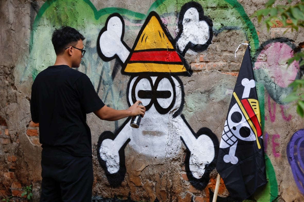 An individual spray paints a skull wearing a yellow triangular hat on a wall