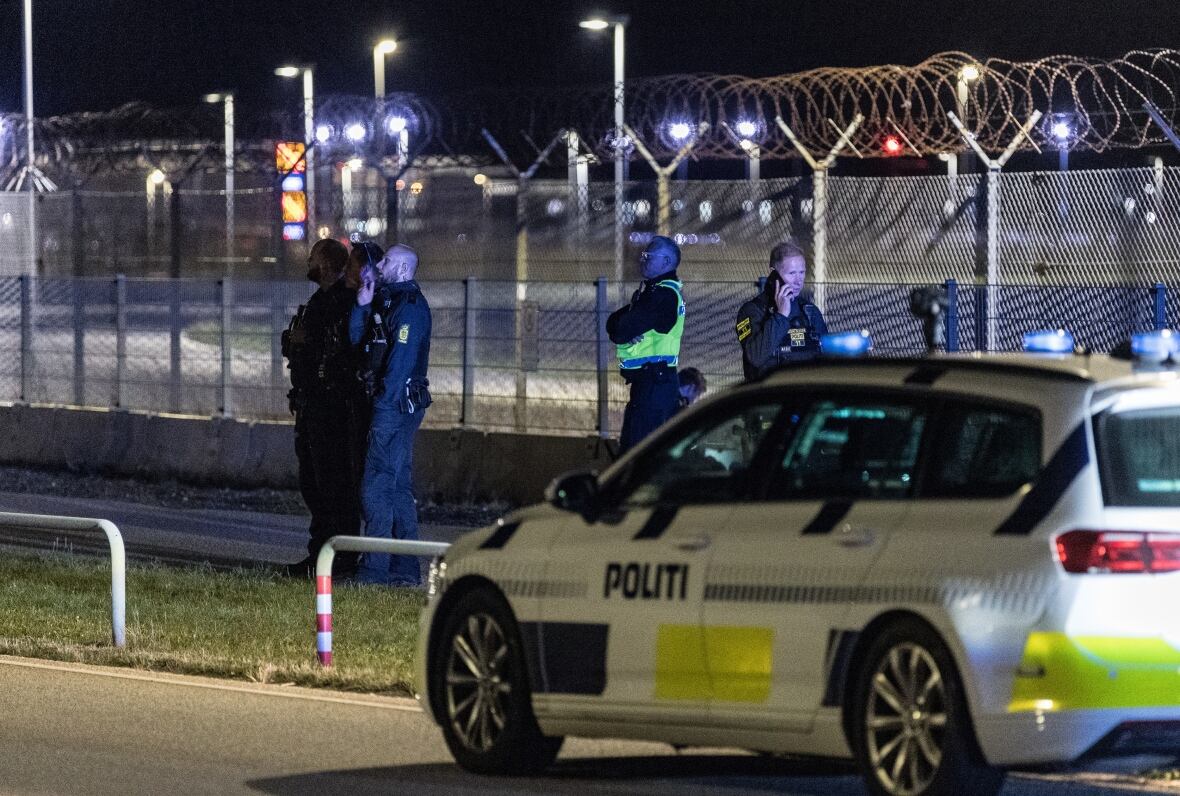 A police vehicle and many officers are displayed near a fence in a night scene, with a low -height building behind them.