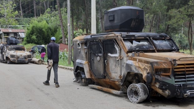 A man walks down a road near a charred military vehicle.