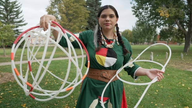 A teenage girl stands in a park holding hoops in the shape of the cradle in one hand and then hoops in the shape of the world in the other. 
