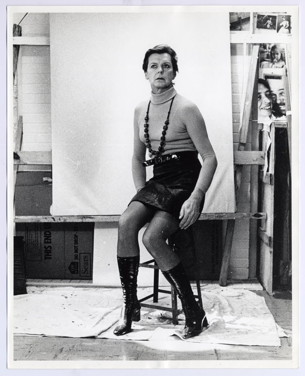 The black-and-white photograph shows a person with short hair, wearing a mini skirt, chunky necklace and tall boots sitting on a stool. 