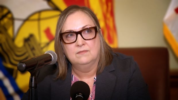 A woman with gray hair wearing dark, square-framed glasses looks off to the side while at a microphone. She's wearing a dark blazer with a pink shirt, and there is a New Brunswick flag behind her.