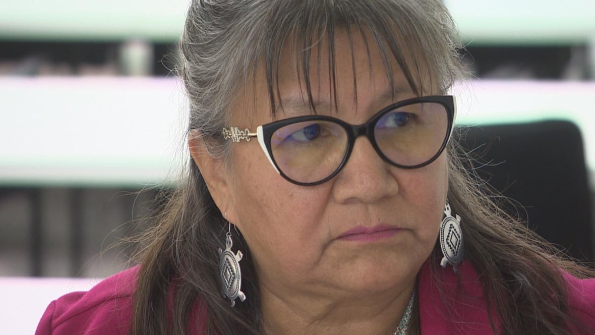 A woman with black-framed glasses, white turtle earrings and a fuchsia blazers looks to the side. 