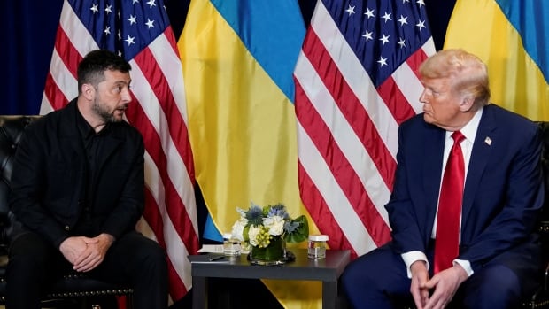 Two men, one in a suit one in dark clothes, sit hacing each other in front of flags