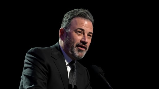 A man in a black suit and tie speaks on stage.