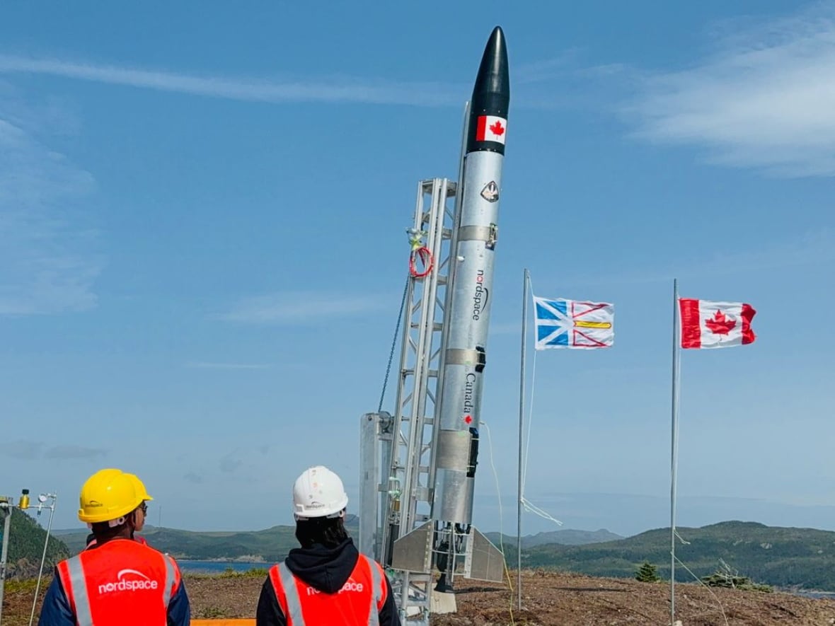 Bild einer Rakete neben zwei Flaggen, während zwei Personen mit Bauhelmen zuschauen.