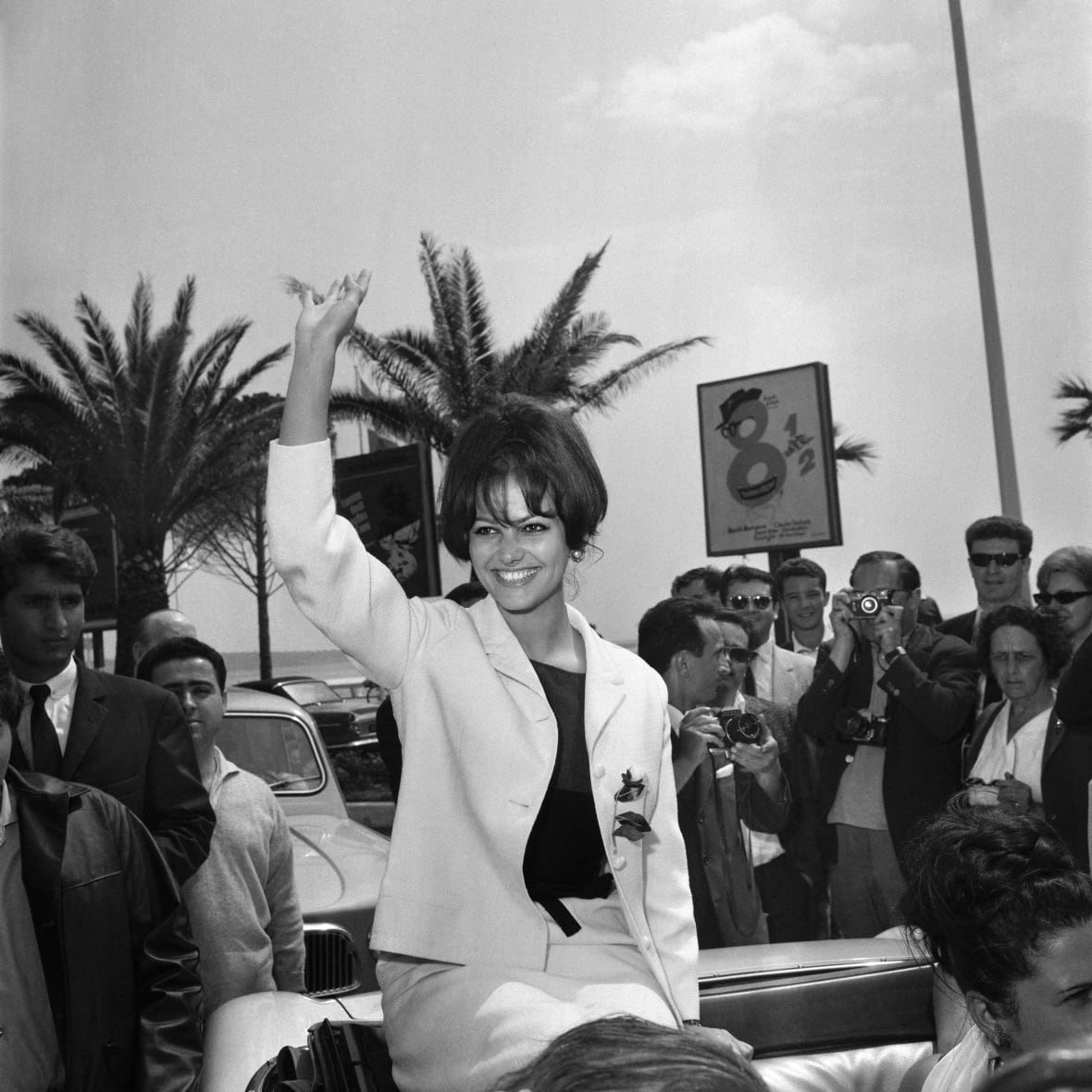 Una donna dai capelli scuri sorride e saluta all'aperto in una fotografia in bianco e nero che sembra avere decenni.