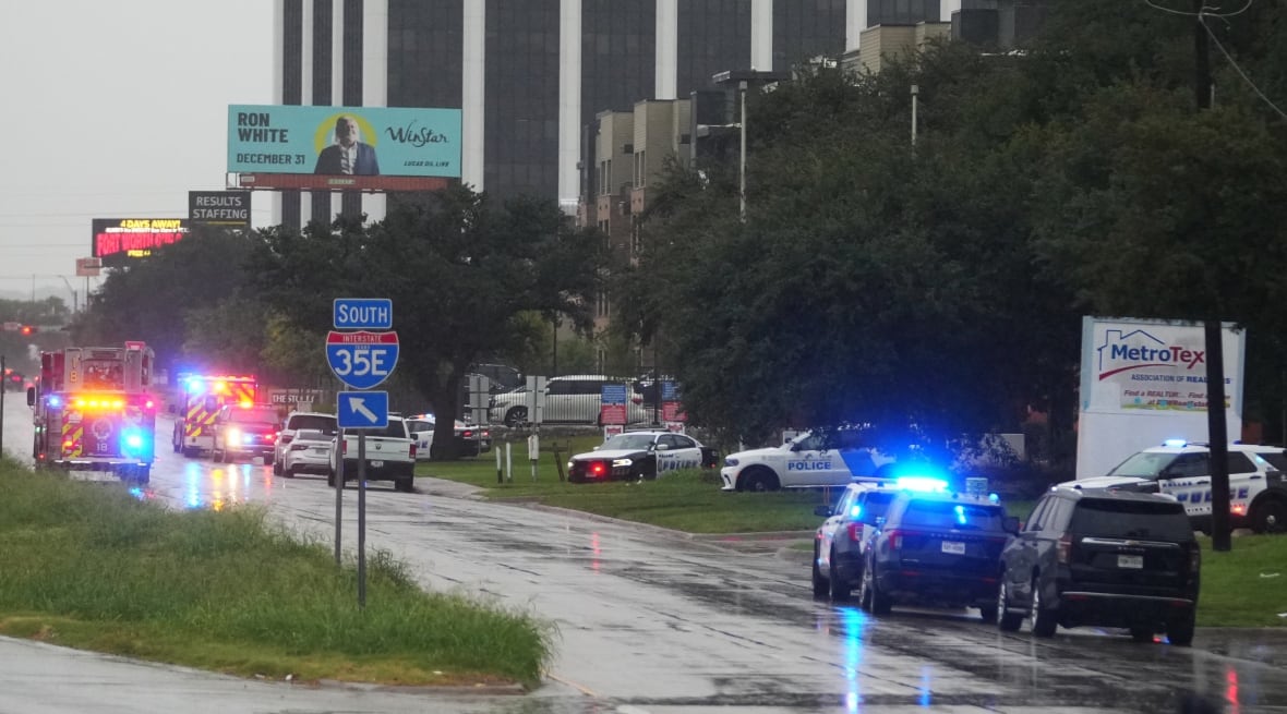 A street bordered by trees with police vehicles and fire trucks on a rainy day. 