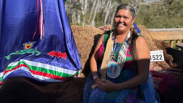 A woman in a silk dress with lace overlay and braided hair sits on a buffalo fur blanket, beside her is a number that she wore at a powwow.