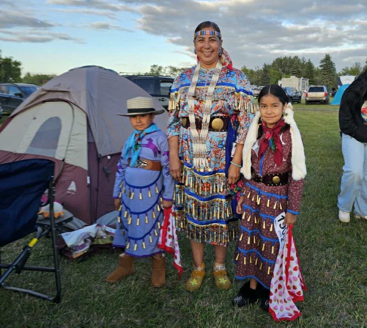 A woman in a dress that has tin cones attached and wears a beaded headbands, holds the hands on each side of two little girls, who are also wearing dresses.