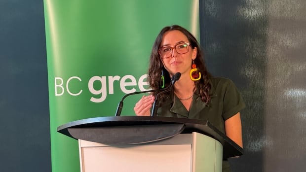 A young woman with curly hair and glasses speaks at a podium with a green 'BC Greens' banner behind her. 