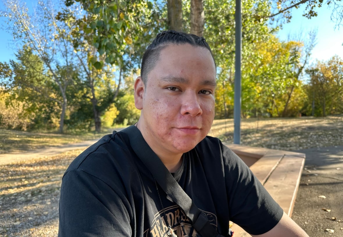 A man with braided-hair sits on a bench in a park with trees in the background.