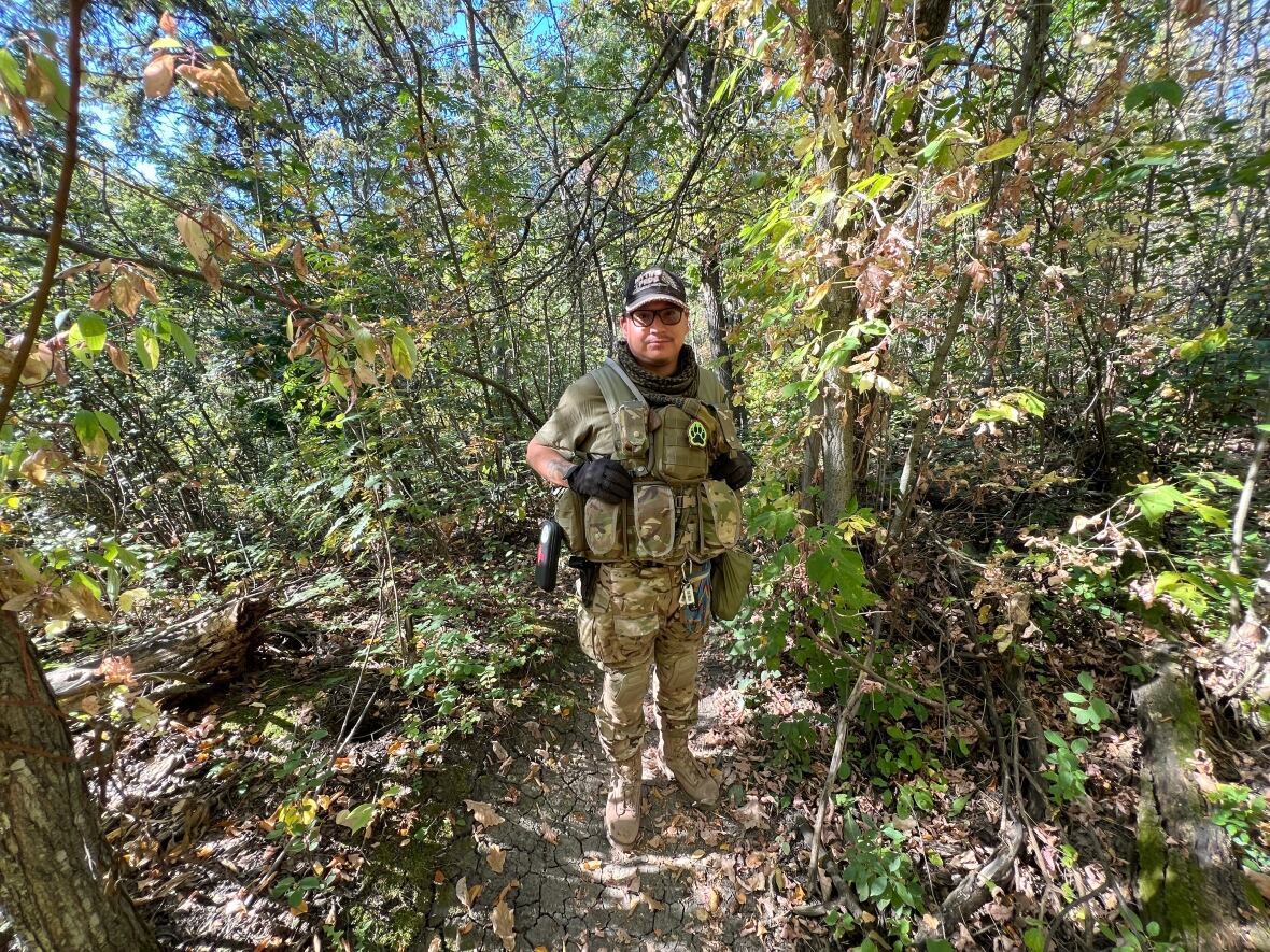 A man dressed in camouflage poses between a thicket of trees. 