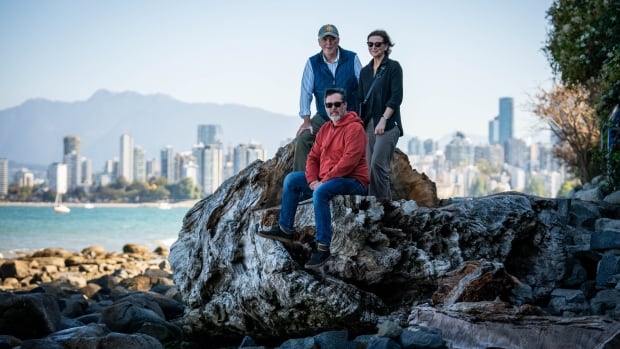 Three people stand on a large, mangled, tree trunk on a beach with a cityscape in the background. 