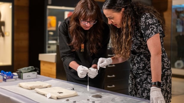 Two women looks at a table of small medallions and rings. They are wearing white gloves.
