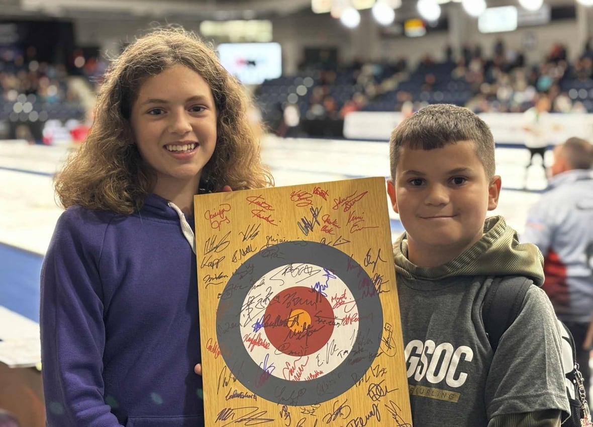 Duas crianças seguram uma prancha de madeira com anéis de curling e autógrafos por toda parte