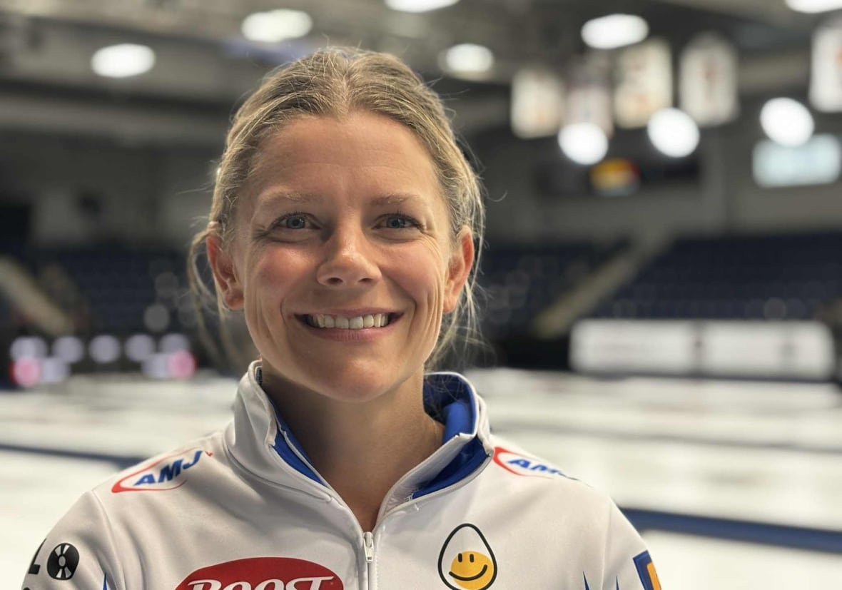 Uma mulher sorri para a câmera. Atrás dela está uma prancha de curling.