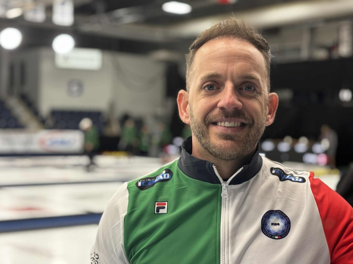 Um homem sorri para a câmera. Atrás dele há uma pista de curling.