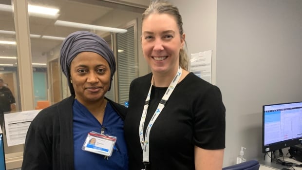 A woman with a head covering and blue scrubs stands arm in arm with a woman with a ponytail and hospital lanyard.