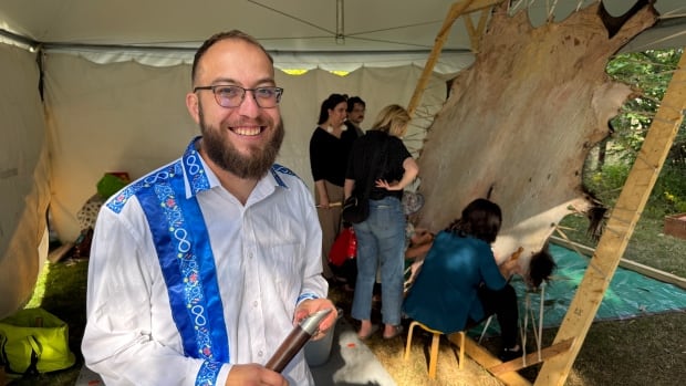 A man in a white shirt with a blue stripe and collar holds a scraping tool in front of a stretched out buffalo hide.