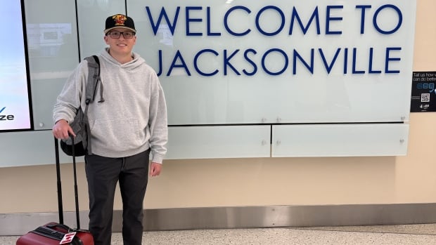 A young man wearing a grey hoodie, black pants, baseball cap and glasses stands with his luggage next to sign that reads, "WELCOME TO JACKSONVILLE."