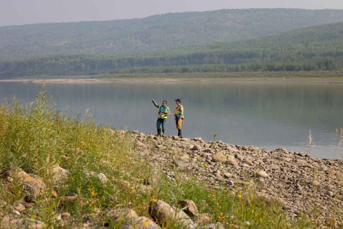Two people in safety vests are pictured by a large body of water surrounded by pine trees.
