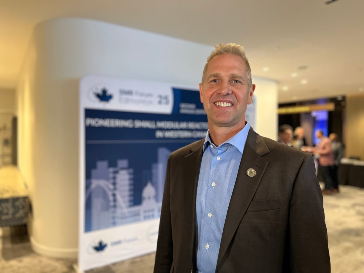 A man wearing a blue shirt and black suit jacket poses in front of a hotel conference ballroom.