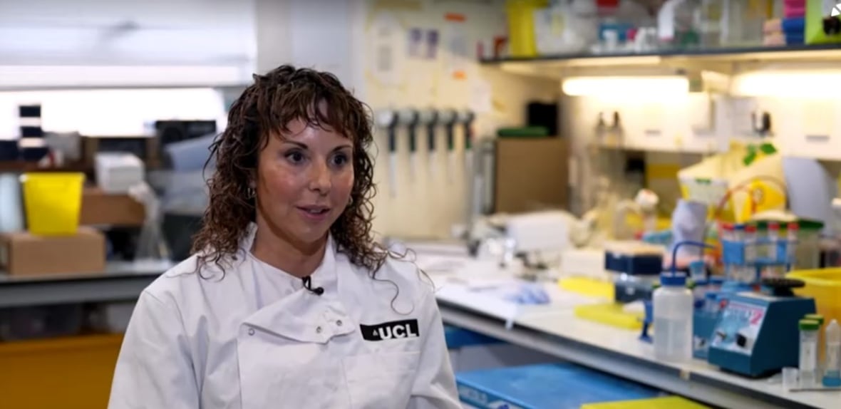 Una donna con i capelli castani e ricci è seduta in un laboratorio e indossa un camice con il logo dell'UCL sulla tasca sinistra.
