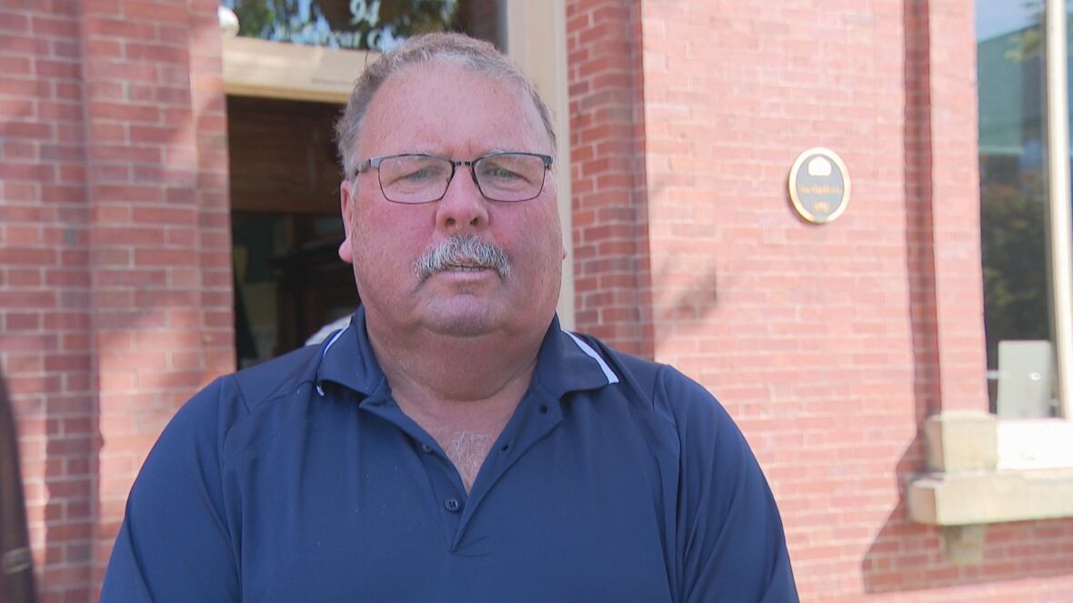 A man wearing a blue golf shirt stands in front of a red brick building
