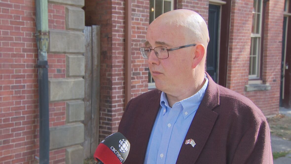 A man wearing a burgundy blazer over a blue shirt stands in front of a red brick building